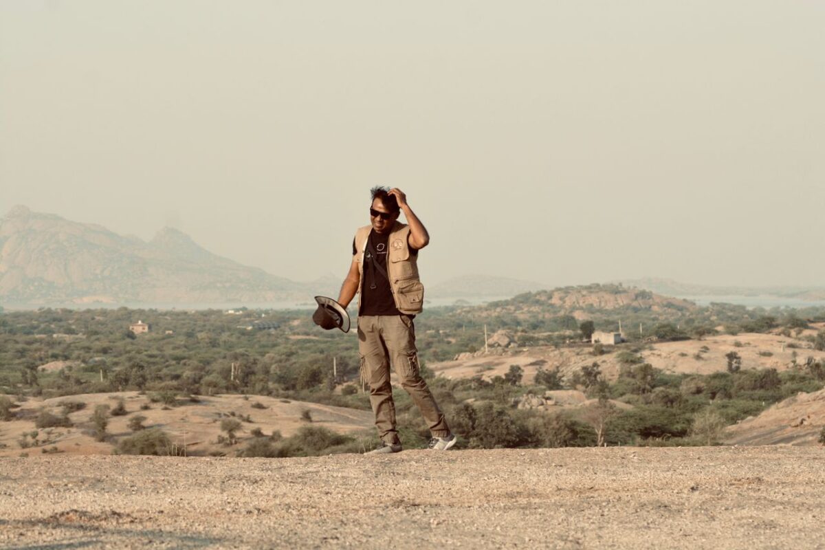 A man in adventure attire stands in a desert with a mountainous background.