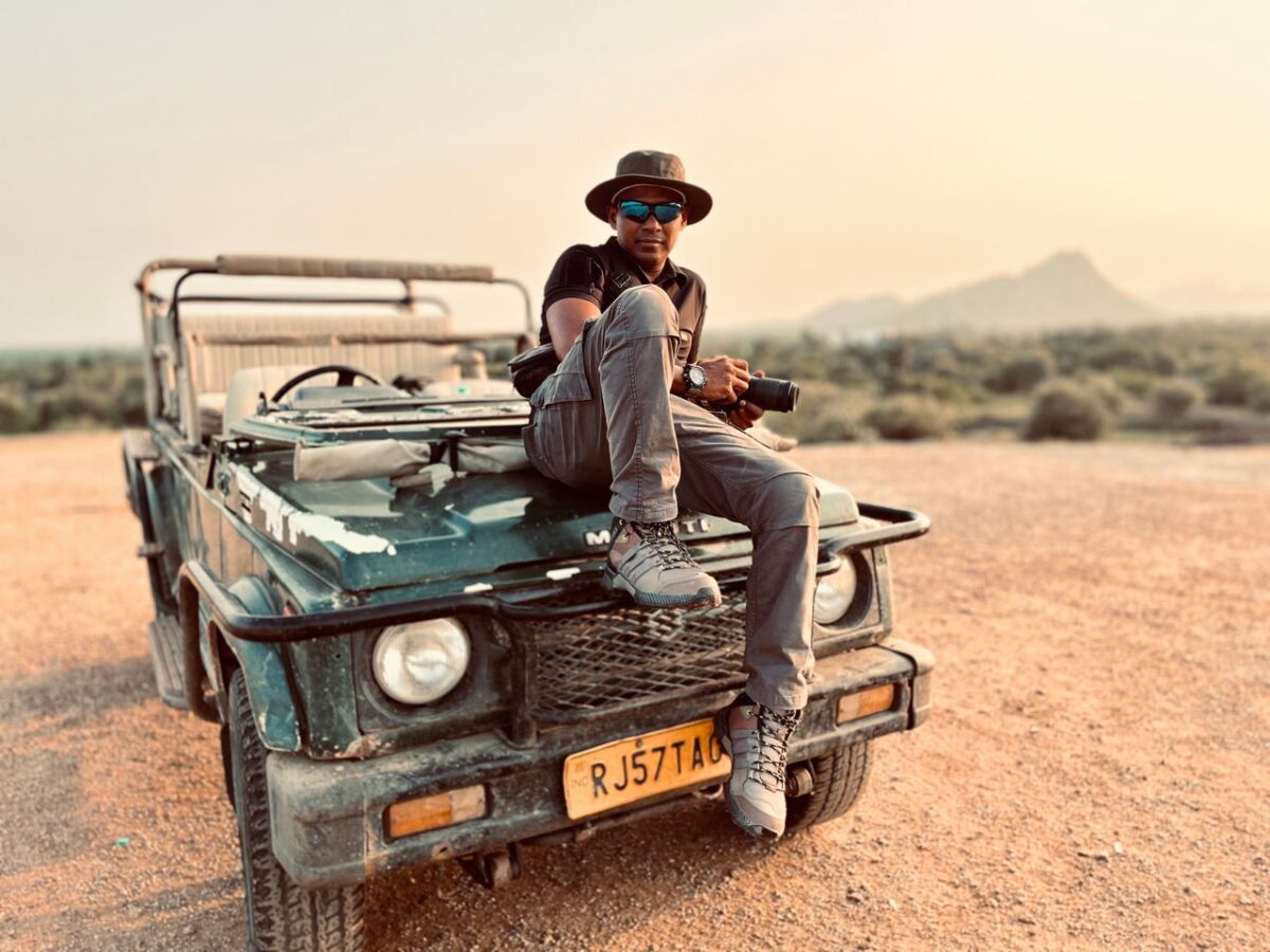 A man wearing a hat and sunglasses sits casually on a jeep in a scenic landscape.