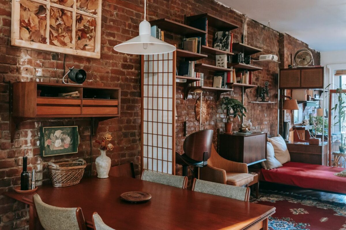 Interior of cozy vintage styled dining room with classic wooden table and chairs placed near weathered brick wall decorated with paintings and bookshelves
